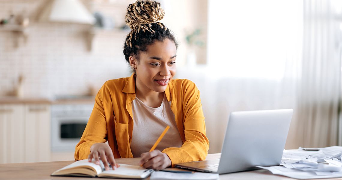 female student studying remotely from home, using a laptop, taking notes on notepad during online lesson